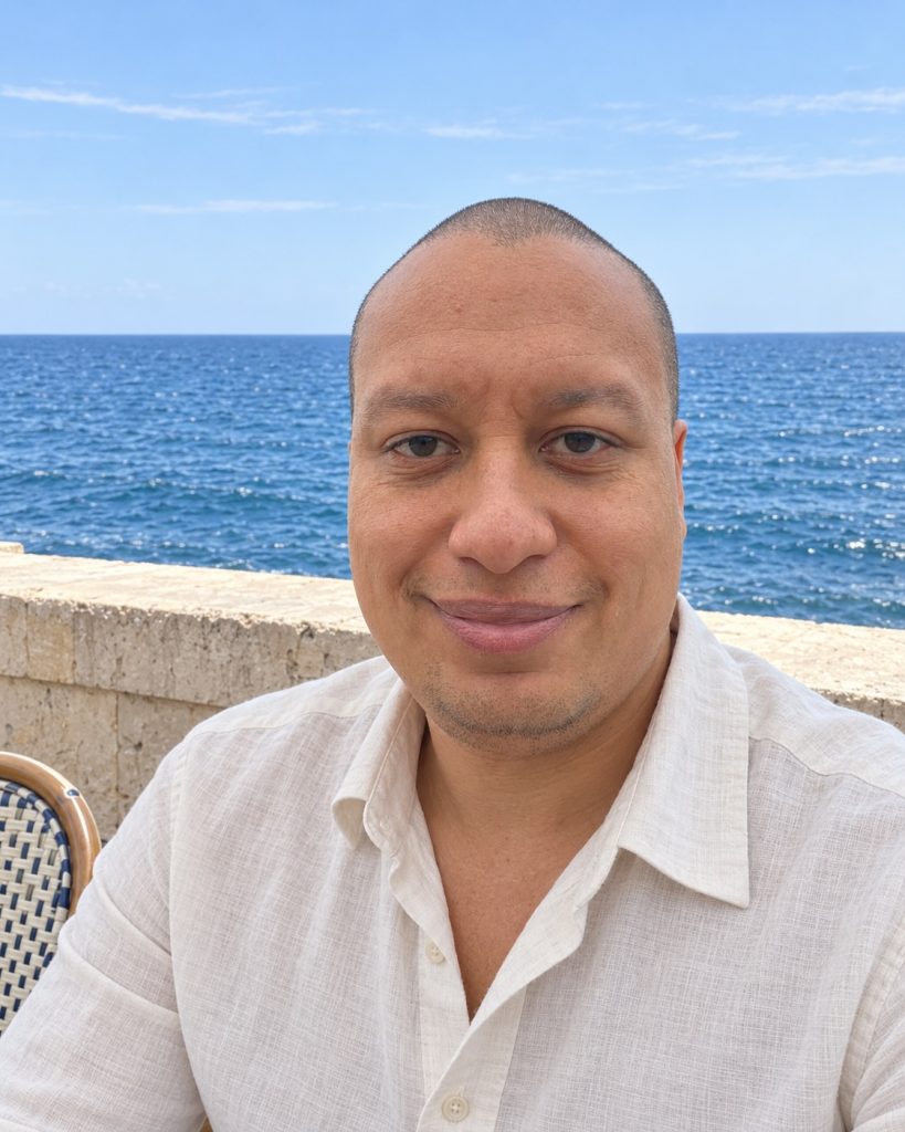 Portrait de Patrick Rastelli, fondateur de L’Identité Abondante, en chemise écrue avec la mer en arrière-plan.